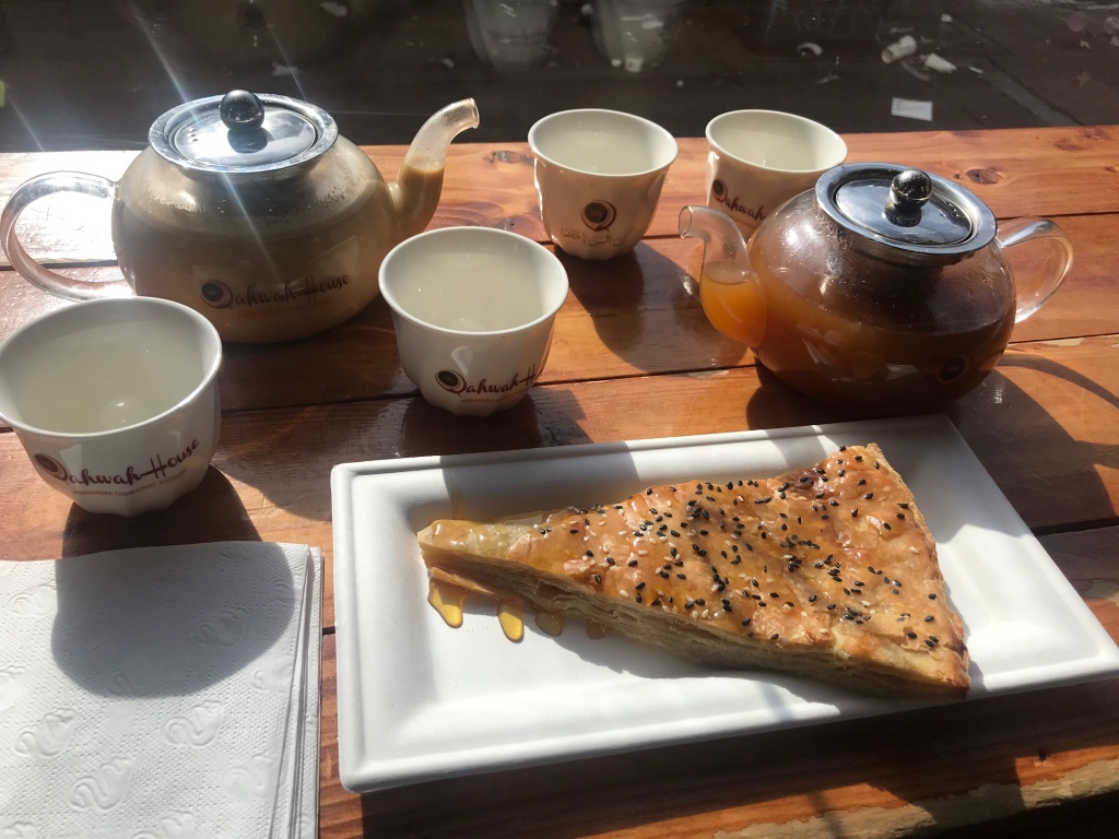 Two coffee pots and four small traditional coffee cups sit on a wooden table, next to a slice of sabaya, a Yemeni pastry.
